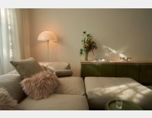 Wohnzimmer mit beiger Sitzecke, dekoriert mit flauschigen Kissen in Pastelltönen. Im Hintergrund stehen eine weiße Stehlampe sowie ein Sideboard mit grünen Schranktüren, darauf eine Vase mit buntem Blumenstrauß und ein Stapel Bücher.