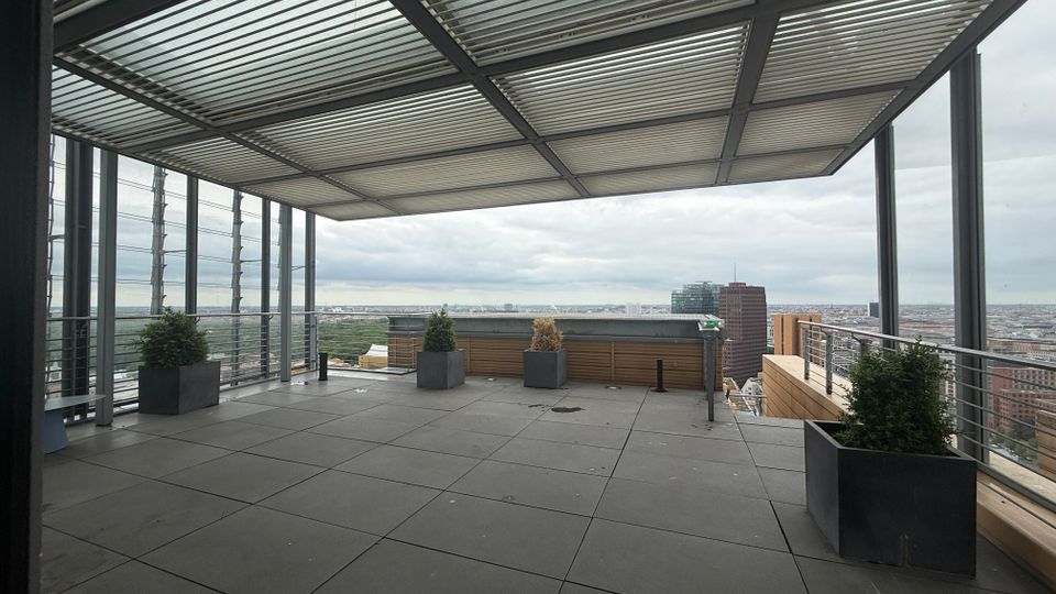 Rooftop terrace with large square floor tiles, a metal slatted canopy overhead, and glass-and-metal railings along the edges. Planters with shrubs line the perimeter, with a wide city skyline view beyond.