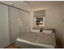 Ein modernes Schlafzimmer mit einem grauen Doppelbett, weißen Bettdecken und einem großen Kleiderschrank mit verspiegelten Schiebetüren auf der linken Seite. An der Wand befindet sich ein Fenster mit einem beigefarbenen Rollo, daneben eine Tischlampe und eine Vase mit Blumen.