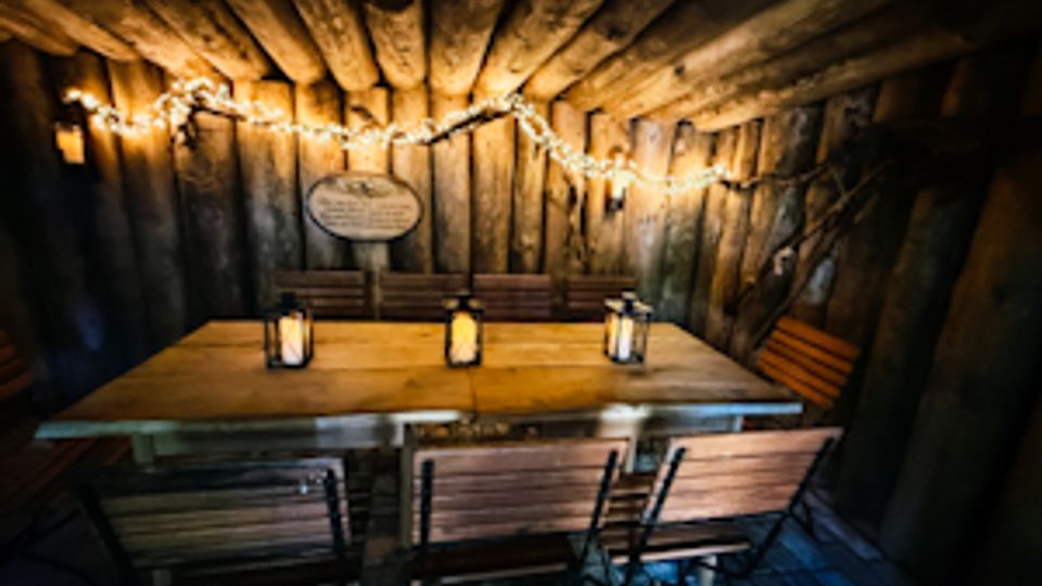 Rustikaler Holzkellerraum mit niedriger Decke aus Baumstämmen und Wänden aus grob behauenen Bohlen, beleuchtet von einer Lichterkette. In der Mitte steht ein großer Holztisch mit mehreren Stühlen und Laternen, der Boden ist mit dunklen Steinfliesen ausgelegt.