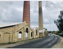 Altes Industriegebäude aus gelbem Backstein mit großen Stichbogenfenstern und zwei hohen Schornsteinen, an einer asphaltierten Straße mit Gehwegen gelegen. Der Himmel ist bewölkt, und im Hintergrund sind Bäume und Straßenschilder zu sehen.