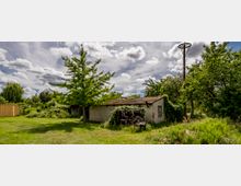 Eine verlassene kleine Hütte mit rotem Ziegeldach, umgeben von Bäumen, Büschen und hohem Gras. Im Vordergrund befindet sich eine Wiese, im Hintergrund ein Strommast und eine leichte Bewölkung am Himmel.