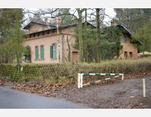Backsteingebäude im ländlichen Stil mit grünen Fensterläden und teilweise bewachsen von Efeu, umgeben von Bäumen und einem niedrigen Holzzaun. Im Vordergrund eine geschlossene Schranke und eine asphaltierte Straße.