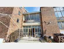 Backsteinfassade eines mehrstöckigen Gebäudes mit großen vergitterten Fenstern und einer offenen Doppeltür im Erdgeschoss. Im Vordergrund liegen Holzstapel und verschiedene Gegenstände wie Eimer und Metallgestänge.