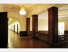 Ein heller Innenraum mit einem hohen Deckengewölbe, einem großen Kronleuchter und Holzverkleidungen. Der Raum hat einen Flur mit Rundbögen, Beleuchtung und einem Geländer aus Holz, das eine offene Galerie umgibt.