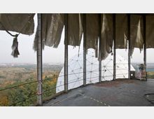 Blick aus einer verlassenen, offenen Struktur mit zerrissenen Stoffpaneelen, die an Metallrahmen hängen. Im Hintergrund ist eine weiße geodätische Kuppel sowie eine waldreiche Landschaft und eine entfernte Stadtansicht sichtbar.