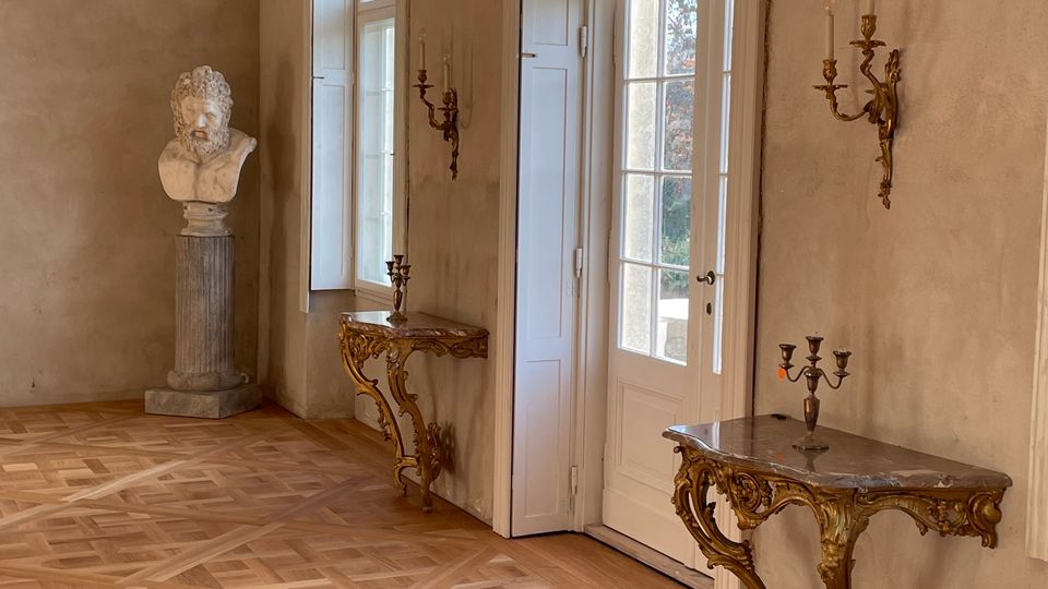 Eleganter Innenraum mit Fischgrät-Parkett, hohen weißen Fenstern und einer verglasten Tür, die viel Tageslicht hereinlässt. An den Wänden hängen vergoldete Wandkerzenhalter; dazu stehen zwei reich verzierte Konsoltische und eine Büste auf einer Säule.