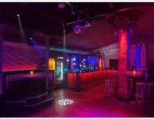 Innenraum einer Bar bzw. eines Clubs mit beleuchteter Theke, Barhockern und Sitznischen entlang der Wände. Farbige Spotlights und eine Discokugel an der Decke, Backsteinpfeiler sowie Getränkekühlschränke prägen die Szene.