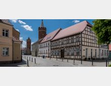 Straßenszene einer Altstadt mit Kopfsteinpflaster, Fachwerkhäusern und roten Ziegeldächern; im Hintergrund stehen zwei mittelalterliche Backsteintürme. Ein Baum spendet Schatten am rechten Bildrand.