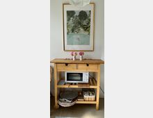 Ein Holztisch mit mehreren Ablageflächen, darauf stehen zwei Vasen mit rosa Blumen. An der Wand dahinter hängt ein gerahmtes Bild mit einer tropischen Landschaft und Schwimmmotiv. Unter dem Tisch befinden sich ein Mikrowellenofen, ein Korb und verschiedene Haushaltsgegenstände.