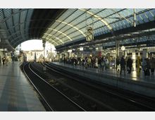 Ein Bahnhof mit moderner Glasdachkonstruktion, mehreren Gleisen und Bahnsteigen. Menschen warten auf den Bahnsteigen, während Züge teilweise sichtbar sind; Uhren und Beschilderung sind deutlich erkennbar.