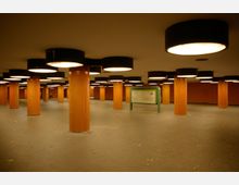 Unterirdischer U-Bahn-Bereich mit orange gefliesten Säulen und runden Deckenleuchten in schwarzen Gehäusen. Im Hintergrund steht eine grüne Hinweistafel mit Wegbeschreibungen.