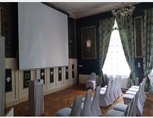 Kleiner Veranstaltungsraum mit weißen Stuhlreihen, einer Projektionsleinwand und einem Rednerpult. Der Raum ist mit dunklen Wänden, dekorativem Stuck, Kronleuchter und großen Fenstern mit gemusterten Vorhängen gestaltet.