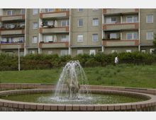 Plattenbau-Wohnhaus mit Balkonen und Zierpflanzen im Vordergrund, begleitet von einer kleinen Grünfläche. Im unteren Bildbereich befindet sich ein runder Brunnen mit einer Wasserfontäne und einer Tierfigur in der Mitte.