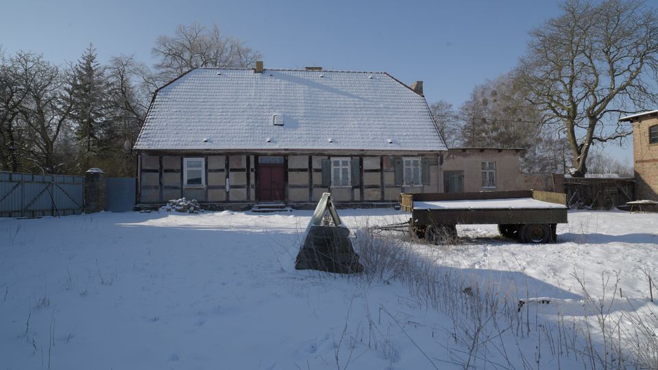 Verschneiter Hof vor einem niedrigen Fachwerkhaus mit großem, schneebedecktem Satteldach und mittiger Eingangstür. Rechts steht ein flacher Anhänger im Schnee; im Hintergrund sind kahle Bäume und Nebengebäude zu sehen.
