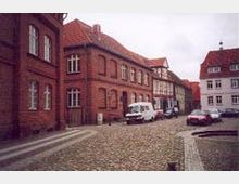 Kopfsteinpflasterstraße mit historischen roten Backsteingebäuden und Fachwerkhäusern, flankiert von parkenden Autos und einem Lieferwagen. Der Himmel ist bewölkt, und im Hintergrund sind weitere Gebäude sichtbar.