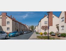 Wohnstraße mit zweigeschossigen Reihenhäusern im traditionellen Stil, verputzt mit roten Klinkerdetails. Links geparkte Autos, rechts kleine Grünbereiche mit Bänken und einem Baum; im Hintergrund moderne Gebäude.