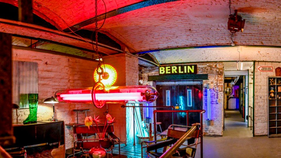 Innenraum eines industrialartigen Untergeschosses mit weiß gestrichenen Ziegelwänden und gewölbter Decke, beleuchtet durch rote und blaue Neonröhren. In der Mitte führt ein Gang zu einer Tür mit dem Schild „BERLIN“, daneben stehen Metallgeländer, Regale und sichtbare Rohrleitungen.