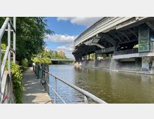 Fußweg entlang eines Kanals mit Geländer und überwachsener Vegetation auf der linken Seite, rechts ein erhöhtes Bahnhofsgebäude mit sichtbarer Stahlkonstruktion und Graffiti an den Betonwänden. Im Hintergrund eine Brücke und Gebäude mit roten Dächern.