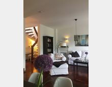 Wohnzimmer mit Holzboden, weißen Polstermöbeln und dezenten Dekorationen. Im Hintergrund befindet sich eine Holztreppe, ein Bücherregal, eine Stehlampe und ein Kamin mit Wandbild darüber.