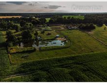 Luftaufnahme eines ländlichen Bauernhofs mit grünen Feldern, einem umfriedeten Teich und mehreren Gebäuden im Hintergrund, umgeben von Bäumen und Wiesen. Im Vordergrund befinden sich landwirtschaftliche Flächen, und eine schmale Straße verläuft am Rand des Geländes.