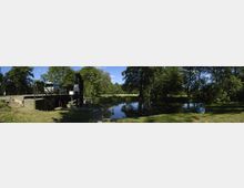 Panoramaaufnahme einer ländlichen Umgebung mit einem kleinen idyllischen Fluss oder Kanal, umgeben von Bäumen und Grünflächen. Im Vordergrund ist eine Schleusenanlage mit Metallgeländern und einem Betonweg erkennbar.