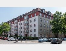 Wohnhaus im Stil der frühen Moderne mit fünf Stockwerken, einem weißen und roten Fassadenmuster sowie markanten horizontalen Streifen. Vor dem Gebäude befinden sich parkende Autos, Bäume und ein Radfahrer auf der Straße.