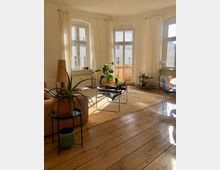 Helles Wohnzimmer mit großen, bogenförmigen Fenstern, weißen Vorhängen und Holzdielenboden. Der Raum ist mit modernen Möbeln, Topfpflanzen und einem kleinen Couchtisch eingerichtet.