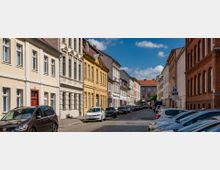 Pflastersteingasse mit geparkten Autos und Reihen historischer Wohngebäude aus verschiedenen Baustilen, darunter Putz- und Backsteinfassaden; im Hintergrund ein größeres Gebäude mit roten Dachziegeln und blauer Himmel mit wenigen Wolken.