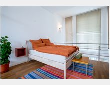 Ein helles Schlafzimmer mit Holzboden, einem Bett mit orangefarbener Bettwäsche, einer Topfpflanze, einem kleinen roten Wandregal sowie großen Fenstern mit beigefarbenen Jalousien. Vor dem Bett liegt ein bunter Teppich, daneben steht ein Hocker mit einer gerollten Decke.