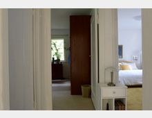 Blick durch einen Flur in eine Wohnung mit zwei angrenzenden Räumen: Links ein Zimmer mit einem Fenster, Pflanzen und einer Kommode, rechts ein Schlafzimmer mit einem Bett, Kissen und hellen Möbeln. Im Flur steht ein kleiner weißer Schrank mit einer Lampe.