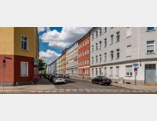 Straßenszene mit mehrstöckigen Wohnhäusern in verschiedenen Farben, die eine enge Straße säumen, auf der geparkte Autos stehen. Der Himmel ist teilweise bewölkt, und an der Straßenecke ist ein gelbes Gebäude mit roter Fassade sichtbar.