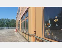 Außenansicht eines langen, orangefarbenen Gebäudes mit großen, rechteckigen Fenstern, teilweise mit Aufklebern bedeckt. Im Vordergrund gepflasterter Boden, links ein leerer Parkplatz und im Hintergrund Bäume sowie ein Zaun.