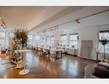 Heller Veranstaltungsraum mit großen Fenstern, durch die Tageslicht einfällt. Der Raum ist mit langen Holztischen und weißen Stühlen eingerichtet, dekoriert mit Pflanzen, Lichtinstallationen und einzelnen Design-Elementen.