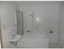 Badezimmer mit weiß gefliesten Wänden und Boden, einer Badewanne mit Duscharmatur und Glastrennwand, einem kleinen Waschbecken mit Ablage sowie einem Lichtschalter und Steckdosen an der Wand.