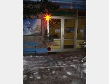 Eingangsbereich einer Galerie mit Glastür und gelber Wand dahinter, beschriftet mit „Galerie“ und „Café“. Der Boden ist verschneit, es steht ein Weihnachtsstern als Dekoration, und links ist ein blaues Gemälde und eine Bank zu sehen.