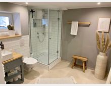 Modernes Badezimmer mit einer Eckdusche mit Glaswänden und weißen Fliesen, einem hängenden WC, einem Waschtisch mit Holzarbeitsplatte und Waschbecken sowie dekorativen Elementen wie einer Vase mit Trockenblumen und einem kleinen Holzstuhl. Beige Fliesenboden und graue Wände vervollständigen das Design.