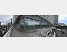 Panoramabild eines modernen Gebäudes mit eckiger Glasfassade und strukturiertem Design, flankiert von einem erhöhten Gehweg mit Geländer. Im Hintergrund ist ein belebter Platz mit Passanten, weiteren Gebäuden sowie parkenden Autos zu sehen, unter einem teils bewölkten Himmel.