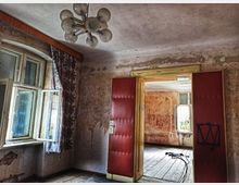 Ein verfallener Innenraum mit abblätternden Wänden, offenem, altem Fenster und einer roten, gepolsterten Tür. Im Vordergrund ein Kronleuchter; durch die Tür ist ein weiteres vernachlässigtes Zimmer mit Holzboden sichtbar.