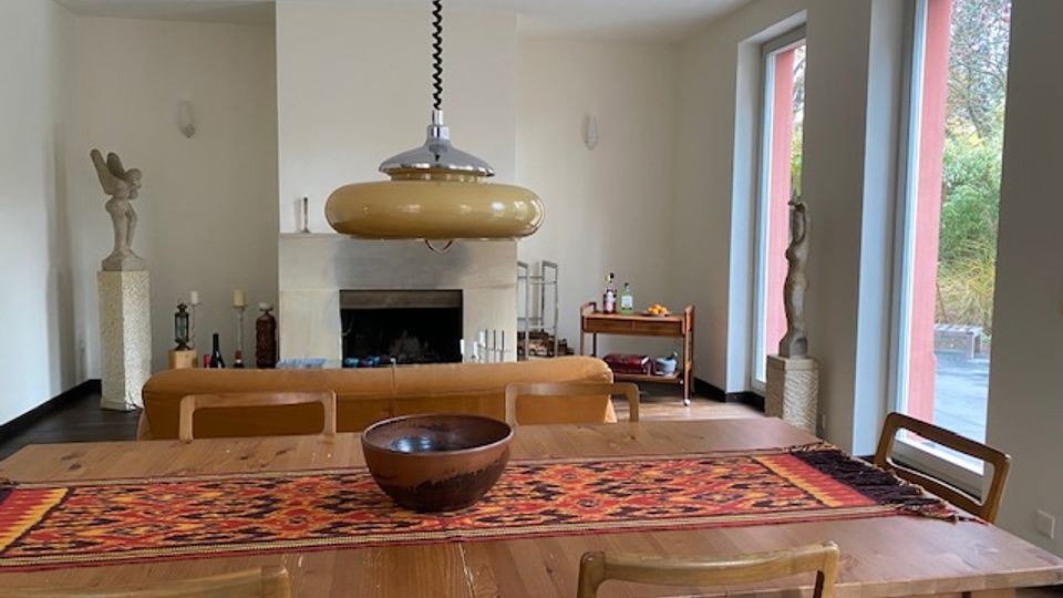 Essbereich in einem hellen Wohnzimmer mit großem Holztisch und Stühlen, gemustertem Tischläufer und hängender Deckenlampe. Im Hintergrund sind ein Kamin, Skulpturen und ein Beistelltisch zu sehen; rechts sorgen hohe Fenster mit Blick ins Grüne für viel Tageslicht.