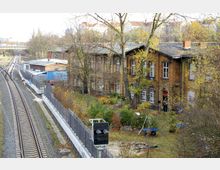 Blick auf ein Bahngelände mit Gleisen und einem historischen Backsteingebäude. Das Gebäude hat große Sprossenfenster und steht in einem Gartenbereich mit Bäumen und Sträuchern; rechts von den Gleisen befinden sich ein Zaun und technische Einrichtungen.