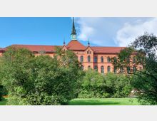 Backsteingebäude mit roten Ziegeldächern und einer grünen Turmspitze mit Uhr, im Vordergrund ein gepflegter Rasen und Büsche. Der Himmel ist teilweise bewölkt.