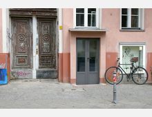 Fassade eines Altbaus mit rosafarbenem Putz und weißen Fenstern. Auf der linken Seite befindet sich eine große, verwitterte Holztür mit Verzierungen und Graffiti, daneben eine moderne graue Glastür sowie ein Schaufenster mit einer Zeichnung und ein angelehntes Fahrrad.