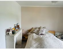 Ein gemütliches Schlafzimmer mit einem Doppelbett, das mit einer hell bestickten Decke und mehreren gemusterten und unifarbenen Kissen ausgestattet ist. Links steht ein weißes dekoratives Möbelstück mit kleinen Ziergegenständen und Fotos, im Hintergrund eine hellbeige Wand.