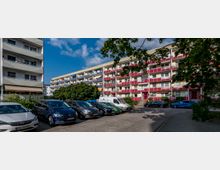 Mehrstöckiges Wohngebäude mit Balkonen und farbigen Fassaden in Beige und Rot, umgeben von geparkten Autos auf einem Parkplatz. Im Vordergrund sind Bäume und Schatten sichtbar, die teilweise den Himmel verdecken.