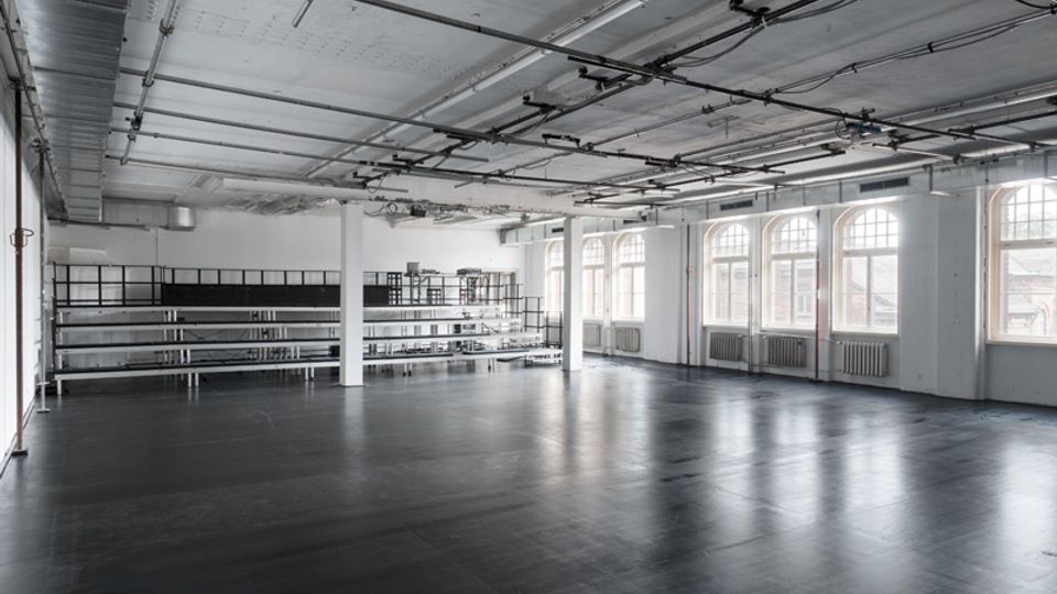 Großer, leerer Loft-Industrieraum mit dunklem Boden, weißen Stützen und sichtbaren Leitungen an der Decke. Entlang einer Seite befinden sich hohe Regalreihen, gegenüber eine Reihe großer Rundbogenfenster mit Heizkörpern darunter.