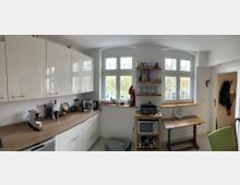 Helle Küche mit weißen Schränken und einer Holzarbeitsplatte, daneben ein Fensterbereich mit drei großen Fenstern, die nach außen auf einen Garten blicken. An der rechten Wand sind Holzregale mit dekorativen Gefäßen, und darunter stehen eine Mikrowelle sowie ein kleiner Holztisch.