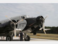 Historisches Flugzeug vom Typ Junkers Ju 52 in silber-schwarzer Lackierung mit der Aufschrift „Lufthansa Berlin-Tempelhof“, geparkt auf einer Rollbahn; im Hintergrund Wald und Himmel, vorne ein Mann in Uniform.