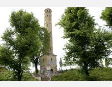 Ein historischer Turm aus gelblichem Stein mit schmalen Fenstern, umgeben von hohen grünen Bäumen. Der Turmsockel ist mit Graffiti bedeckt, und eine Treppe führt hinauf durch eine parkähnliche Umgebung.