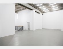 Minimalistischer, leerer Innenraum mit weißen Wänden, Betonboden und einer offenen Betondecke mit mehreren langen, schmalen Leuchtstofflampen. Im Hintergrund befindet sich eine Treppe mit schwarzem Geländer und Holzhandlauf.